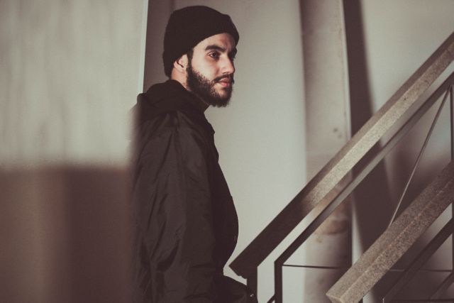 Young man wearing a beanie and dark clothing standing beside an indoor staircase in low light. He has a serious and pensive expression. Suitable for use in fashion editorials, urban lifestyle magazines, modern photoshoots, and products targeting young adults.