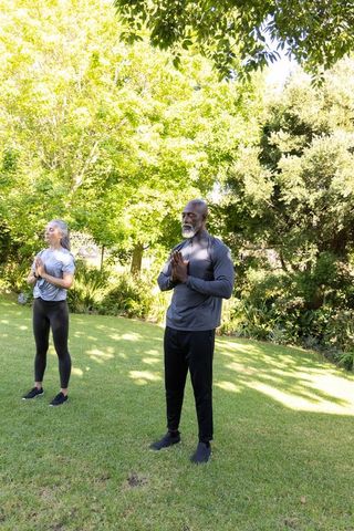 Mature couple practicing yoga in sunlit garden under trees, standing meditation pose