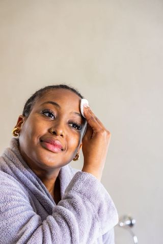African american woman cleansing face with cotton pad in plush bathrobe at vanity