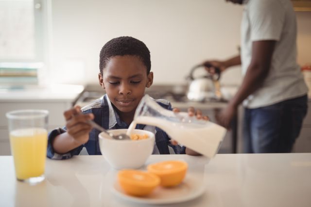 Boy pouring milk into a bowl of cereal in a modern kitchen, with a parent in the background. Ideal for illustrating family morning routines, healthy eating habits, and domestic life. Suitable for use in advertisements, parenting blogs, and nutritional guides.