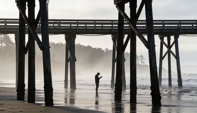 Silhouette standing under wooden pier holding phone on misty coastal beach