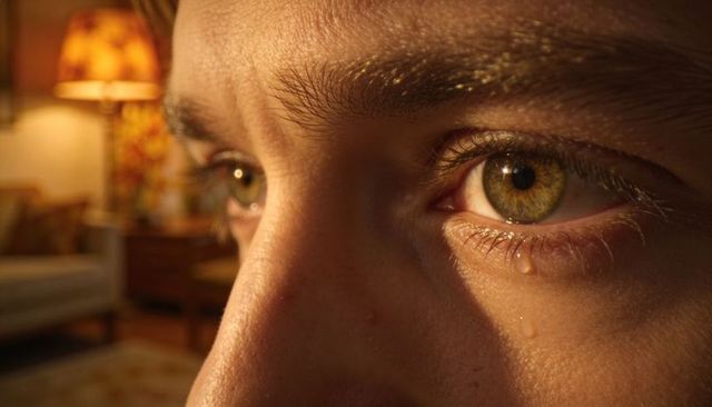 Tearful hazel-eyed man gazing in warm lamp-lit living room, close-up emotional portrait