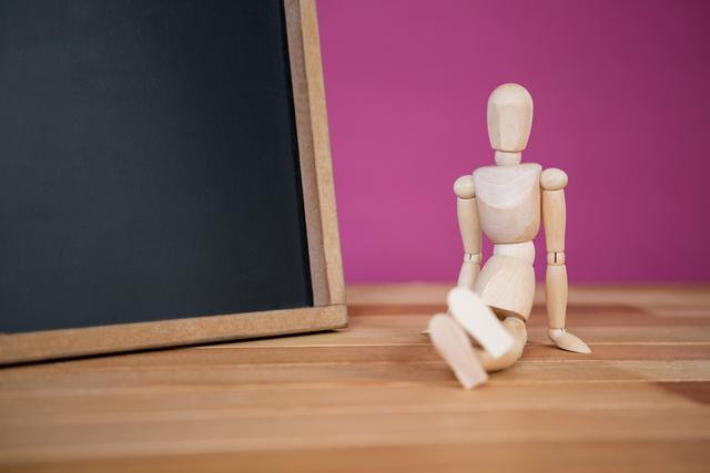 Wooden figurine sitting near an empty chalkboard on a wooden floor with a pink background. Ideal for educational themes, creative projects, art classes, teaching materials, and school-related content. Can be used to represent concepts of learning, creativity, and classroom settings.