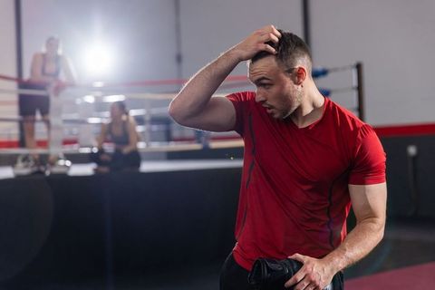Male boxer catching breath after sparring wearing red shirt holding glove under spotlight