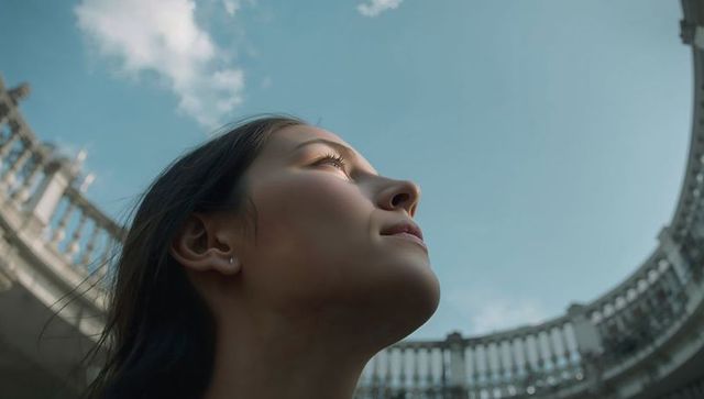 Young woman gazing upward, sunlit profile against blue sky with curved stone balustrade