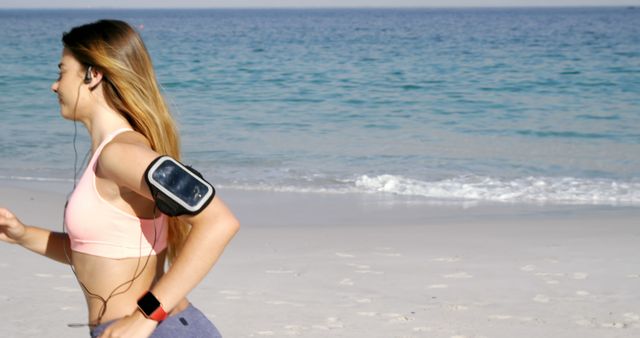 A young Caucasian woman is jogging on a beach, wearing a pink sports top and using a smartphone armband, with copy space. Her outdoor exercise routine showcases a healthy lifestyle and the enjoyment of fitness activities in a natural setting.