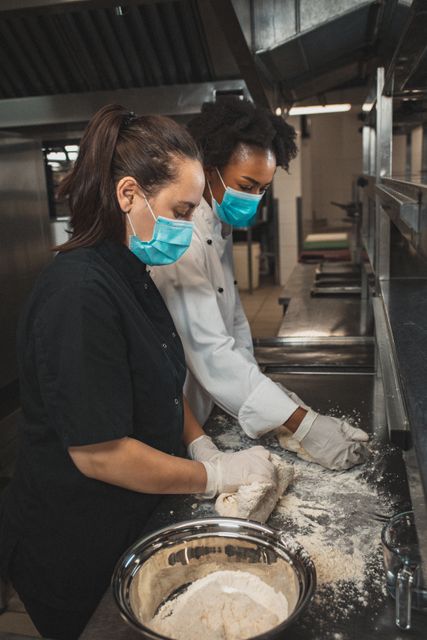 Diverse two female professional chef wearing masks kneading floured dough. working in a busy restaurant kitchen during coronavirus covid 19 pandemic.