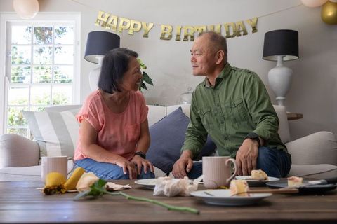 Asian senior couple celebrating cozy home birthday sharing cake and coffee on sofa
