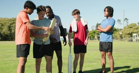 Teen cricket team inspecting bat during coach-led training on sunny grass field