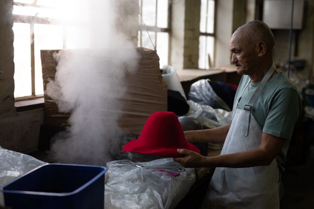 Side view of a senior biracial man holding a red unfinished hat over steam in the workshop at a hat factory.
