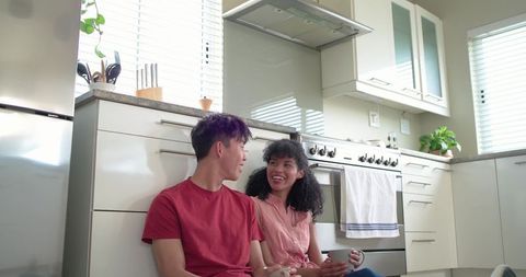 Sunlit african american woman and asian man sitting on kitchen floor holding mugs