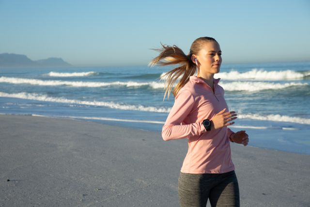 Caucasian woman jogging on a beach in sportswear, enjoying a sunny day. Ideal for promoting healthy lifestyle, fitness, outdoor activities, and wellness. Can be used in advertisements for sportswear, fitness programs, or travel destinations.