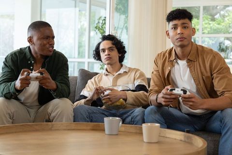 Multiracial young men playing console games on couch sharing snacks in bright living room
