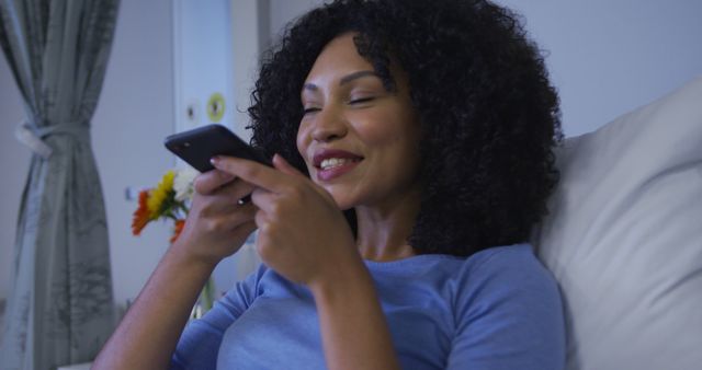 Young woman with curly hair holding and texting on smartphone while sitting in a comfortable setting. She is smiling, indicating a pleasant conversation. Ideal for concepts related to communication, technology use, leisure time, home comfort, and youthful lifestyle.