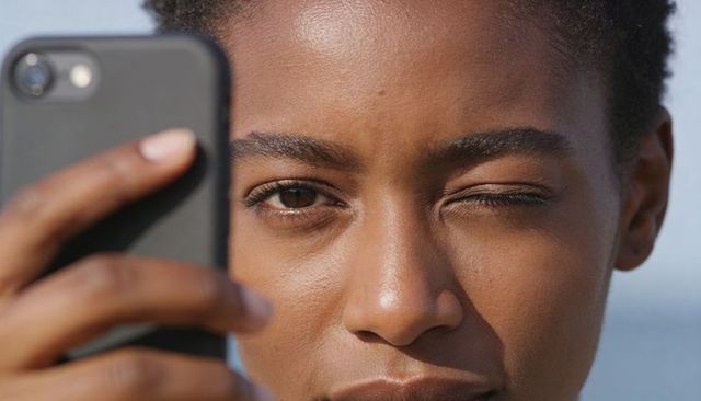 Winking woman holding smartphone close to face under blue sky sunlit natural portrait