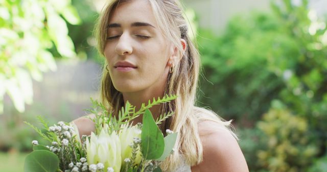 A woman stands in a lush garden, holding a bouquet of fresh flowers close to her face with eyes closed, enjoying the scent and relaxing in nature. Perfect for themes related to tranquility, self-care, floral arrangements, gardening, mindfulness, and wellness.