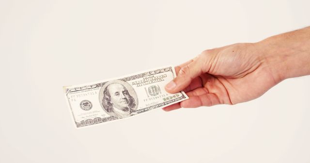 A Caucasian hand is holding a hundred-dollar bill against a white background, with copy space. The image represents financial transactions, monetary exchange, or concepts of wealth and payment.