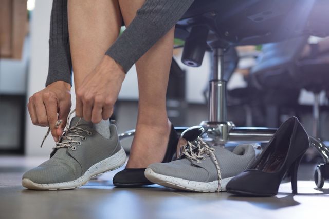 Businesswoman sitting in office, changing from high heels to comfortable sneakers. Ideal for illustrating work-life balance, office comfort, professional attire, and the transition between formal and casual footwear in a workplace setting.