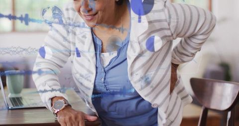 Senior Indian woman leaning on table with back pain at home using laptop, wearing cardigan