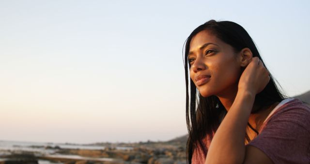 Biracial woman with long black hair enjoying seaside view. She's wearing pink top and looks thoughtful, with a calm expression