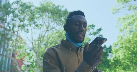 Man speaking into smartphone while walking sunny urban park wearing mask under chin