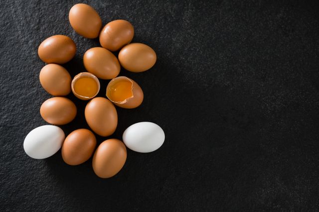 Brown and white eggs arranged on a dark background, with one egg cracked open revealing the yolk. Ideal for use in culinary blogs, cooking websites, nutrition articles, and food-related advertisements.