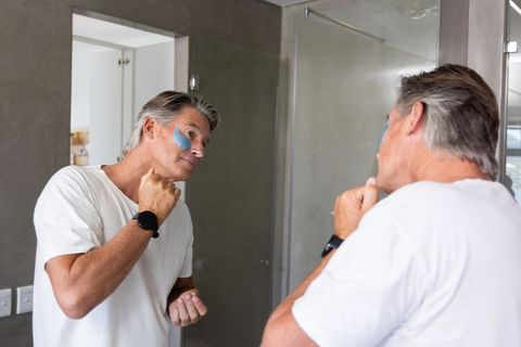 Mature man applying blue under-eye patch at bathroom mirror wearing smartwatch and white tee