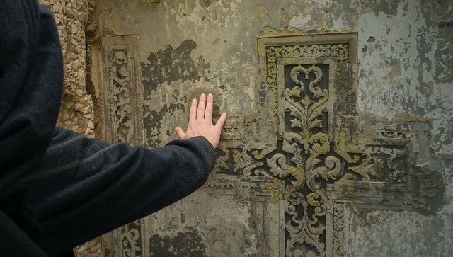 Resting hand touching weathered ornate cross on chapel wall with mossy plaster texture