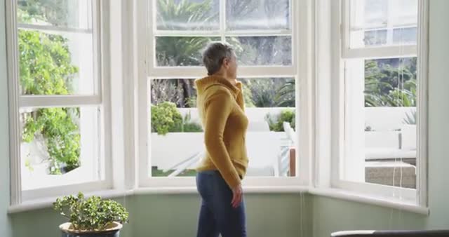 An elderly woman with short grey hair in a cowl neck sweater stands by the window, engaged in a phone conversation. Sunlight illuminates her, combining warmth with the modern environment visible outside. Suitable for use in lifestyle contexts or representing senior communication strategies and leisure.