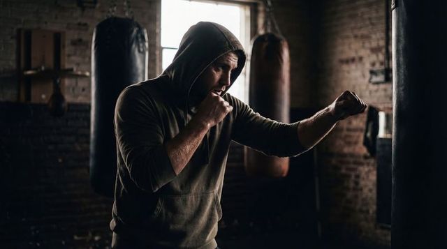 Hooded boxer practicing jab in gritty backlit gym with heavy bags, chains and brickwork
