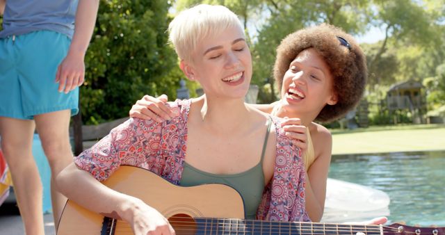 Friends having fun on sunny day by the pool with one playing guitar and others laughing. Perfect for concepts related to summer activities, friendship, leisure time, outdoor enjoyment, and carefree days. Great for advertisements focusing on music, summer vacations, and social gatherings.