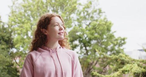 Redhead woman standing on terrace wearing pink hoodie looking hopeful amid lush green park