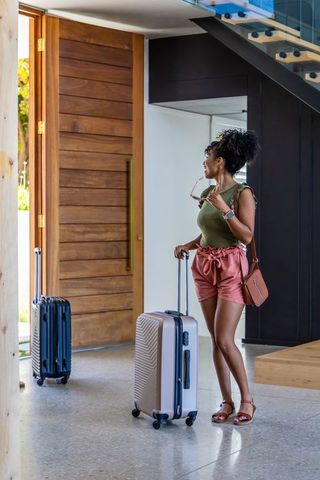 African american woman standing in modern foyer holding carry-on suitcase and sunglasses