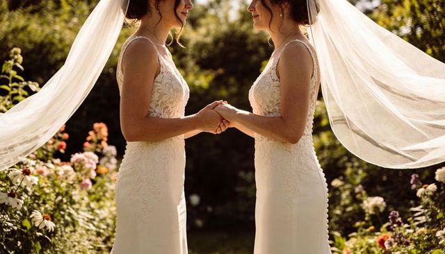 Two brides holding hands in garden wedding, lace gowns and flowing veils at golden hour