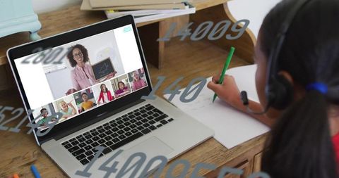 Student writing during virtual classroom on laptop wearing headset using green pen at home desk
