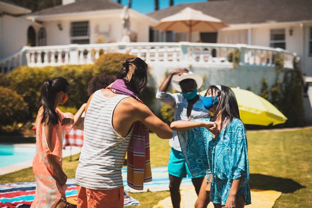Diverse group of happy friends wearing face masks greeting touching elbows at pool party. hanging out and relaxing outdoors in summer during coronavirus covid 19 pandemic.