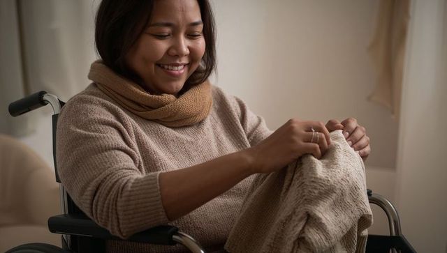 Smiling asian woman in wheelchair folding beige knitted sweater at home, cozy independence