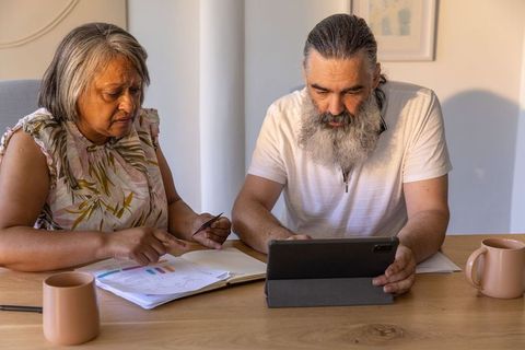 Mature couple reviewing finances with tablet and charts at sunlit dining table, minimalist