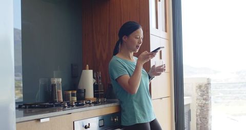 Young woman leaning on modern kitchen counter texting on smartphone holding glass of water