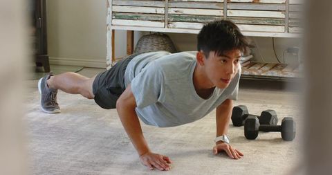 Asian man performing push-up at home living room with dumbbells and rustic cabinet