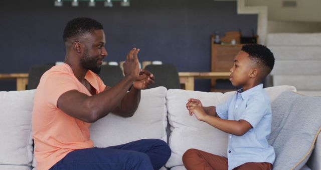 Father and son engaged in sign language communication while sitting on cozy couch at home. Perfect for illustrating family bonding, educational support, parenting, and effective communication. Can be used in articles about family values, sign language learning, parent-child relationships, and modern parenting practices.