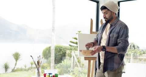 Indian artist wearing beanie mixing paint at easel by window with coastal view
