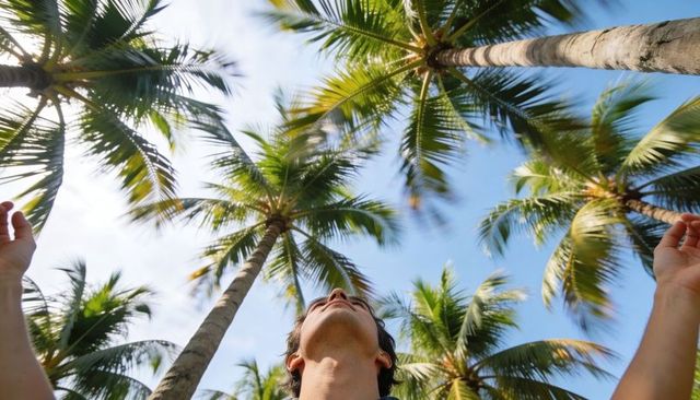 Shirtless man looking up while raising arms under coconut palms and blue tropical sky