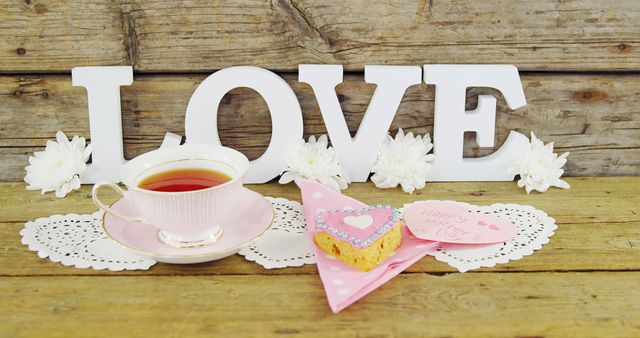 A cup of tea is placed next to a decorative sign spelling LOVE and white flowers, with a heart-shaped cookie and a note suggesting a Valentine's Day theme. The arrangement conveys a cozy and romantic atmosphere, perfect for celebrating love and affection.