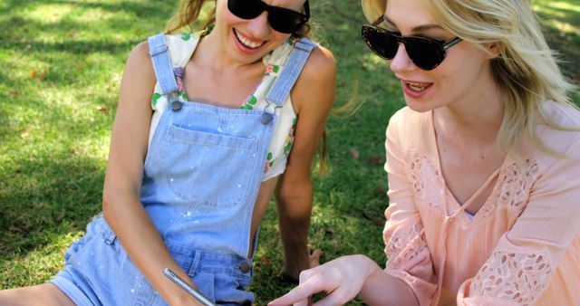 Two young Caucasian women are enjoying a sunny day outdoors, sitting on the grass and looking at a smartphone together, with copy space. Their casual attire and sunglasses suggest a relaxed, leisurely atmosphere.