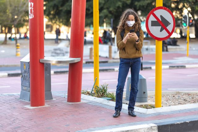 Caucasian woman wearing a face mask standing on a city street during the day, using a smartphone. Ideal for illustrating urban life during the pandemic, health and safety measures, or technology use in public spaces.
