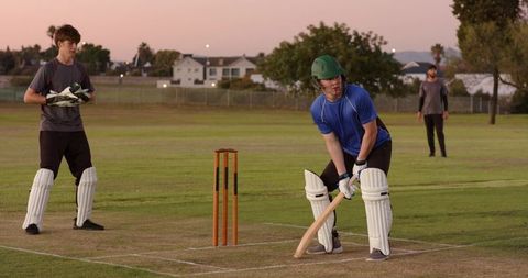 Young cricketer batting at dusk on suburban oval with wicketkeeper and wooden stumps