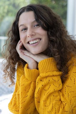 Young woman with curly hair smiling while wearing mustard-yellow knitted sweater by window