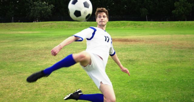 Young athlete wearing white and blue uniform kicking soccer ball on grass field. Useful for sports advertisements, fitness promotions, youth sports programs, and motivational content about teamwork and athleticism.