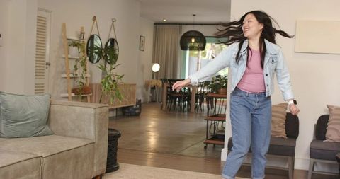 Asian woman spinning joyfully in sunlit modern living room with plants wearing denim jacket